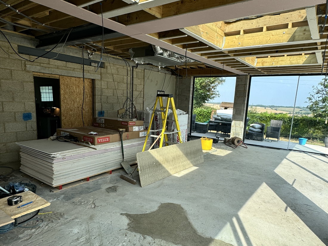 Inside the extension during construction, with steel beams and timber joists overhead, rooflight openings formed and the glazing openings looking out over fields, by LD&C Homes