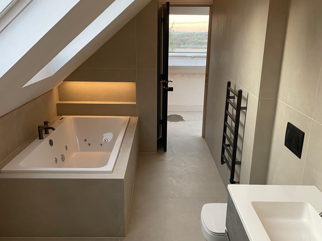 The finished bathroom in the loft conversion, with a bath set under the slope, a wall hung vanity unit and large format tiling, by LD&C Homes