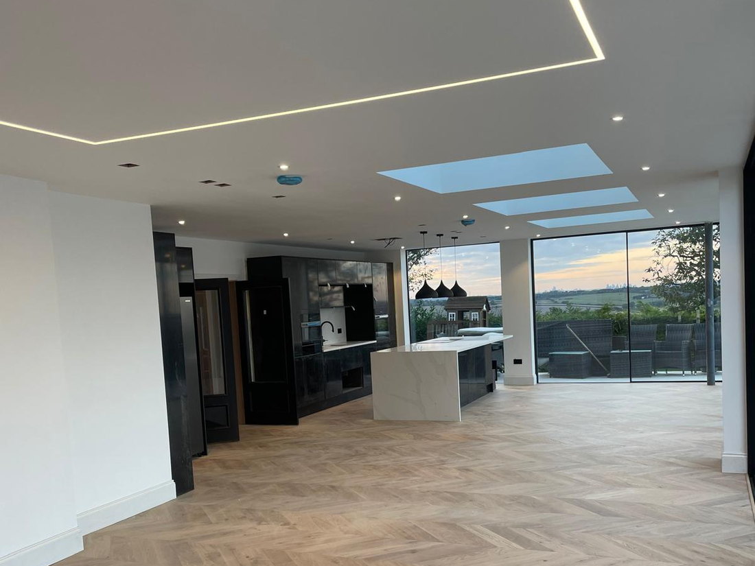 The finished open plan ground floor at dusk, with chevron oak flooring, rooflights, recessed lighting and full height glazing across the rear, by LD&C Homes