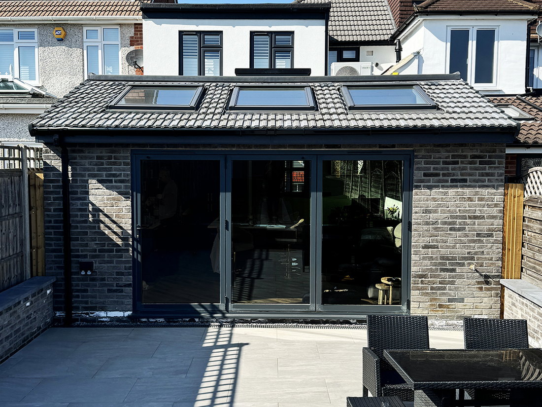 The finished single storey rear extension in grey stock brick, with a mono pitched tiled roof, three roof windows and large format bifold doors onto a porcelain patio, by LD&C Homes