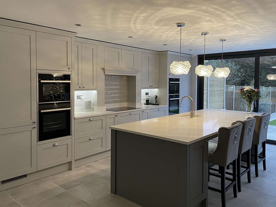 An open plan kitchen extension in the evening, with cream shaker units, a stone island with seating and full width sliding doors onto the garden, built by LD&C Homes