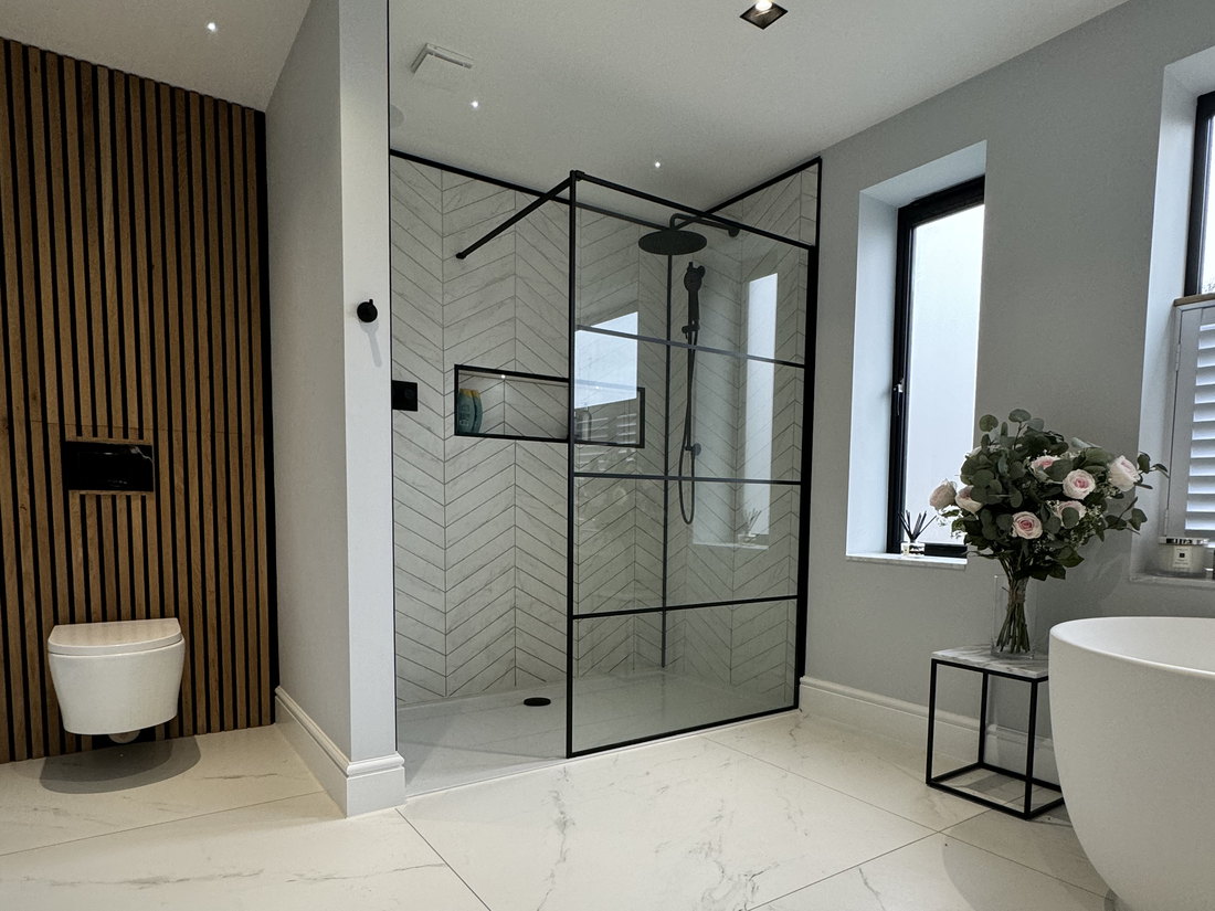 A finished bathroom with a walk in shower behind black framed glazing, herringbone tiling, a fluted timber panel wall, a freestanding bath and large format marble effect floor tiles, by LD&C Homes