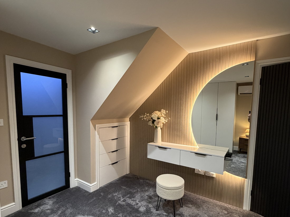 A finished bedroom with a fitted dressing area, fluted panelling with a backlit round mirror, built in drawers under the slope and a wall hung vanity, by LD&C Homes